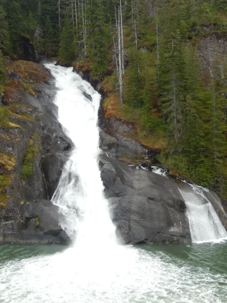 A massive, rushing glacial waterfall cascading down dark rock faces into the ocean during an alaskan-cruise-adventure.