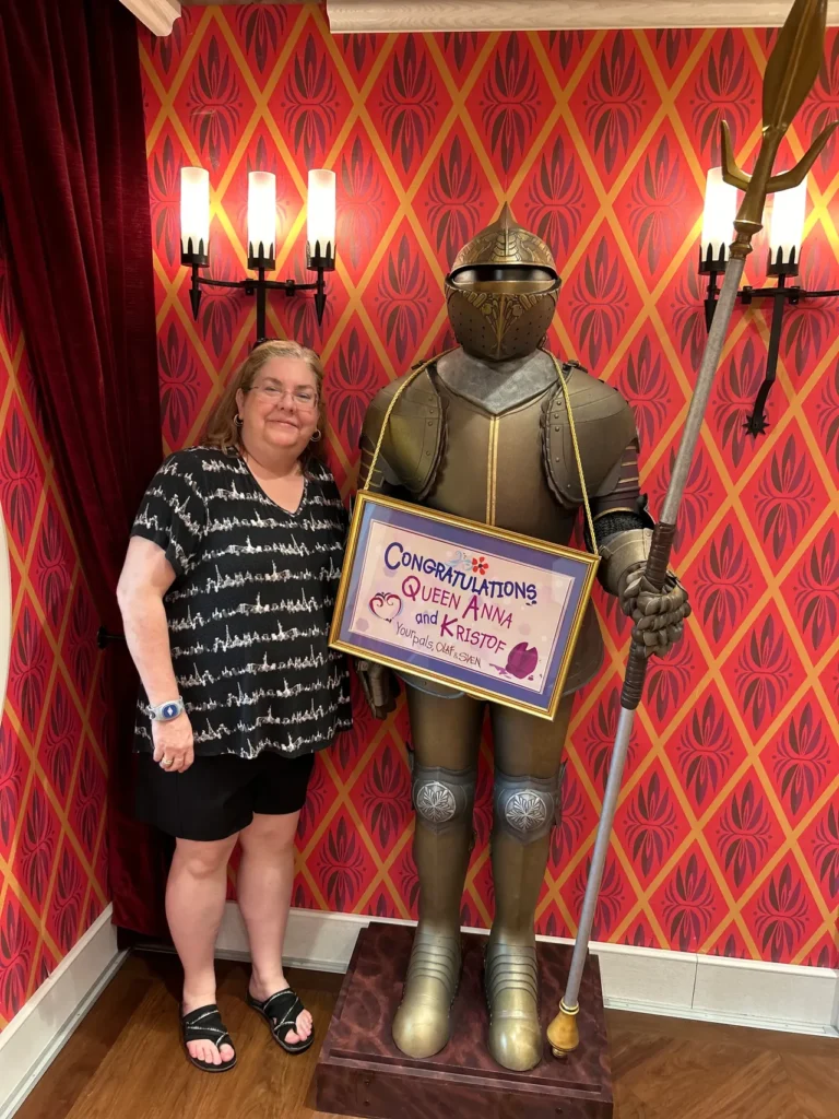 A guest smiling next to a historic suit of armor holding a Queen Anna and Kristof congratulatory sign at the Arendelle restaurant featured in our Disney Wish dining guide.