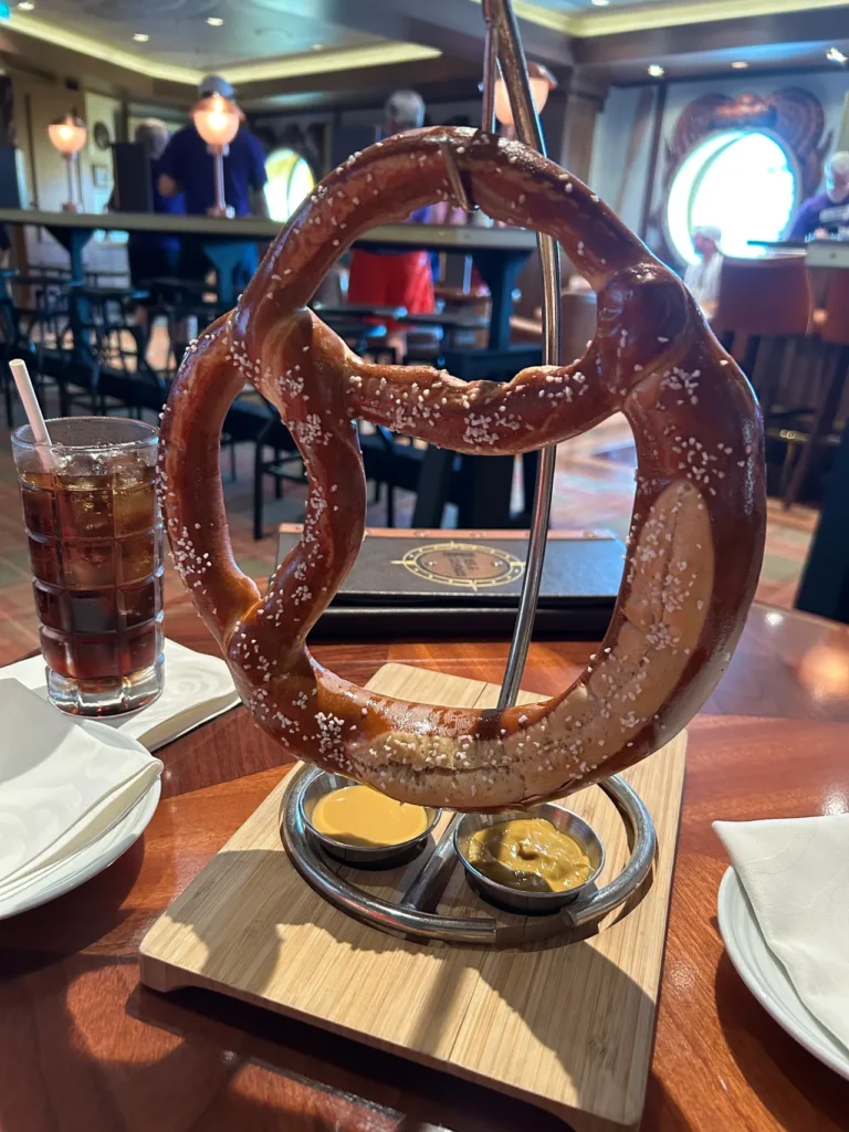 A giant traditional Bavarian pretzel hanging from a stand with cheese and mustard dipping sauces at the Keg & Compass pub from our Disney Wish dining guide.