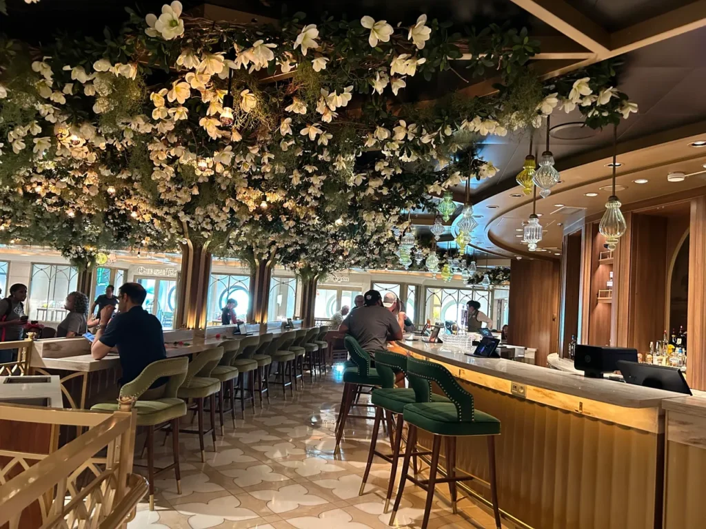 Guests sitting at the elegant bar under a canopy of white flowers and magnolia leaves inside The Bayou lounge featured in our Disney Wish dining guide.