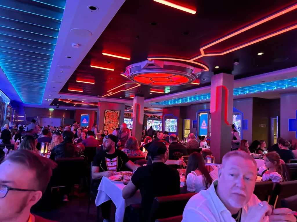 Families enjoying their dinner under the vibrant red and blue neon ceiling lights in the Worlds of Marvel dining room on the Disney Wish.