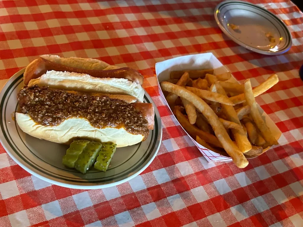 The famous Tony Packo hot dog. Made to your specifications, this one features one plain and one with their special chili sauce. The hot dog platter comes with a cup of chili, fries and some spicy pickles. A classic Tony Packo’s Toledo hot dog platter featuring one chili dog, one plain hot dog, french fries, and sweet hot pickles on a red checkered tablecloth.