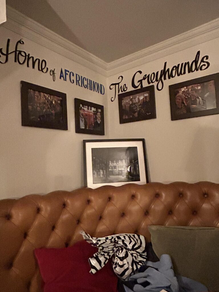 AFC Richmond is not a real football team. But you'll find plenty of team memorabilia throughout the pub. This corner booth has become a mini-shrine to the show with photos of your favorite characters. A cozy corner booth decorated with framed show photos and team branding, a highlight of the real Ted Lasso pub in Richmond.
