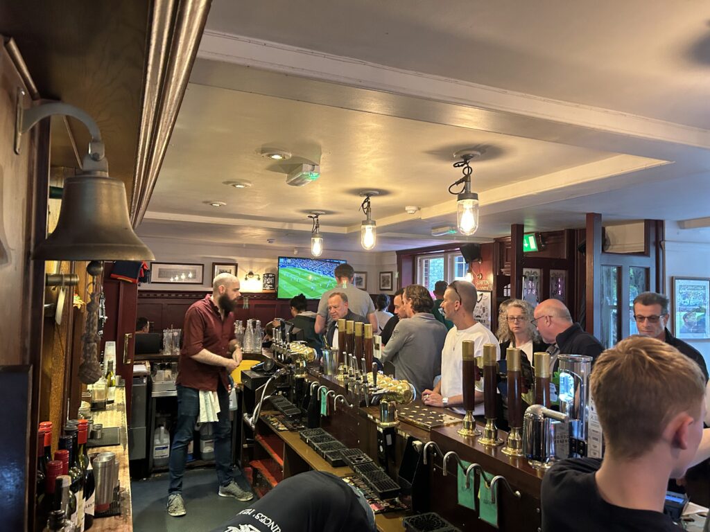 A classic pub design, the bar at Prince's Head greets you just inside the front door. Expect a small crowd, especially on the weekend. Also expect to see football on the TV screen. Patrons gathered around the historic wooden counter at the main bar inside The Prince's Head, the real Ted Lasso pub in Richmond.