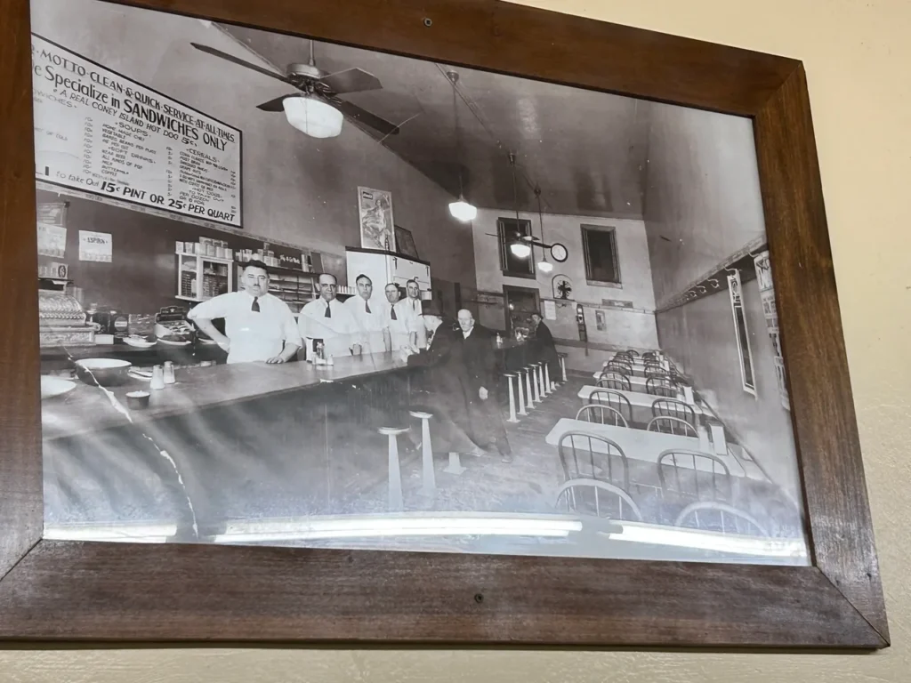 The walls of Coney Island are filled with historic photos of the establishment. The staff dresses more casually now, which is the only thing that has changed from this photo. A historic black-and-white photo display showcasing the original counter and employees at America's oldest hot dog stand.