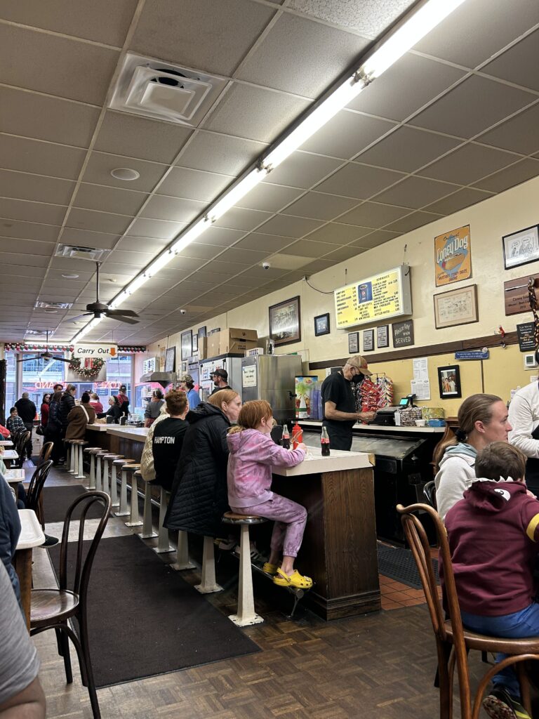 Enjoy your coney dog at the counter or at one of the tables at Coney Island in downtown Fort Wayne. The menu, located behind the counter, is largely unchanged over the past 100+ years. Customers dining at the modern day counter inside Fort Wayne's Coney Island, featured in our America's oldest hot dog stand restaurant guide.