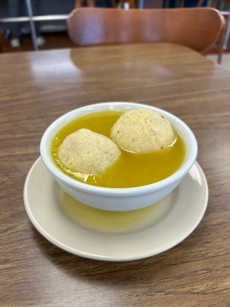 Hand rolled matzo balls, served in a delicious chicken broth. Shapiro’s has this soup along with about a half dozen others available daily. A white ceramic bowl of classic matzo ball soup featuring two large, hand-rolled matzo balls in a golden chicken broth at Shapiro's Deli Indianapolis.