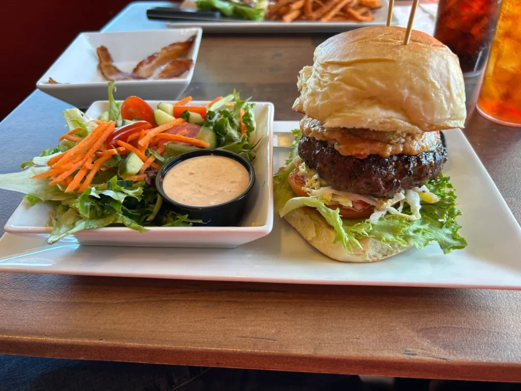 The half tank burger from GAS is a half pound burger with house made pimento cheese, slaw, applewood smoked bacon, fried pickles, lettuce, tomato and onion. A massive half tank burger topped with house-made pimento cheese, bacon, and fried pickles at GAS Full Service Restaurant, a highlight of our St. Augustine restaurants guide.