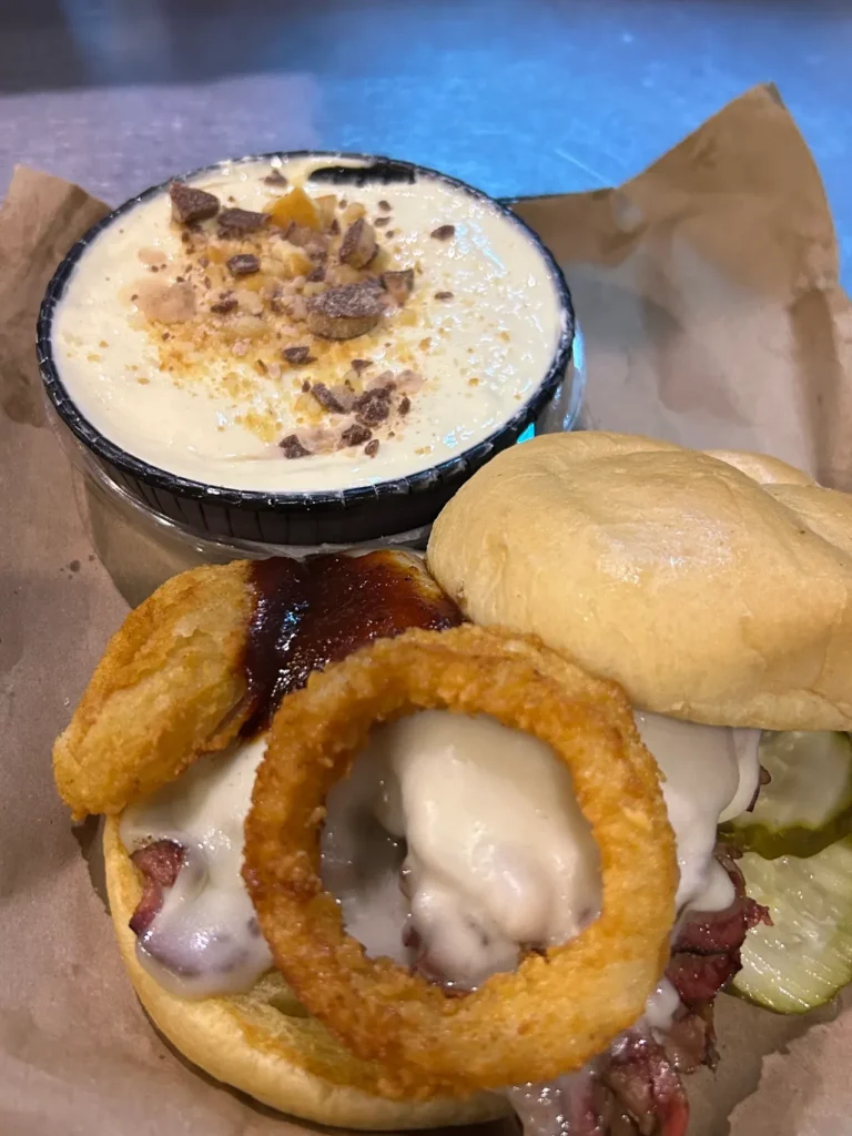 The world famous Z-Man sandwich from Joe's Kansas City BBQ. Topped with smoked provolone cheese and an onion ring, this beef brisket sandwich is legendary. Also pictured is the amazing banana pudding dessert topped with Heath bar. The iconic Z-Man sandwich featuring sliced beef brisket, provolone cheese, and onion rings on a toasted Kaiser roll from a Kansas City BBQ tour.