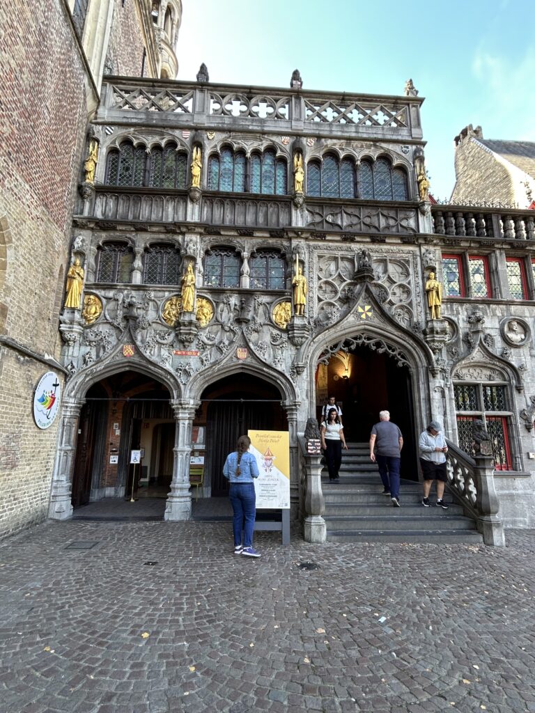 Tucked in downtown Bruges is the Basilica of the Holy Blood. The church claims that they have a piece of cloth stained with the blood of Jesus Christ. The ornate dark grey and gold Gothic facade of the Basilica of the Holy Blood during a Bruges day trip.