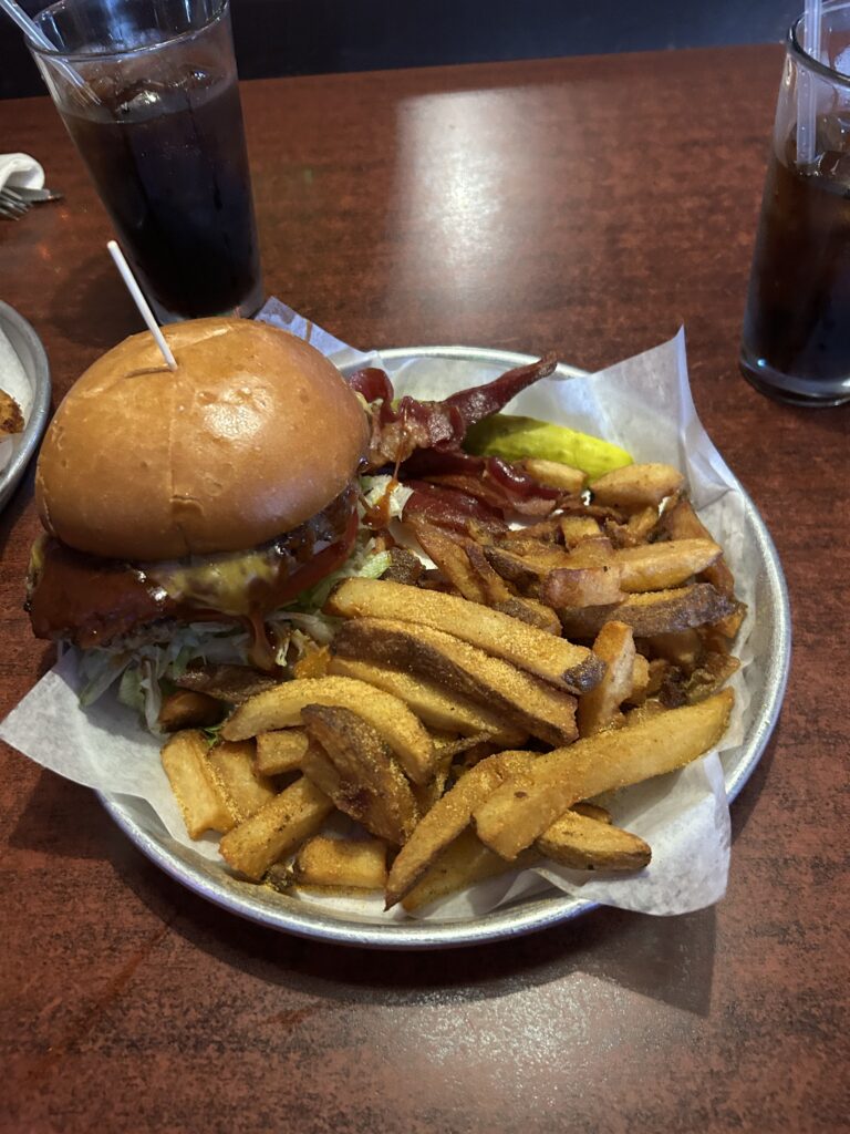 The Western Burger with hand-cut fries from the Thurman Cafe. Highlights include the fresh burger, never frozen and delivered each day, along with tasty thick-cut bacon and a delightful bbq sauce. A close-up view of the savory Western Burger served with thick-cut bacon and fries at the Thurman Cafe Columbus Ohio.