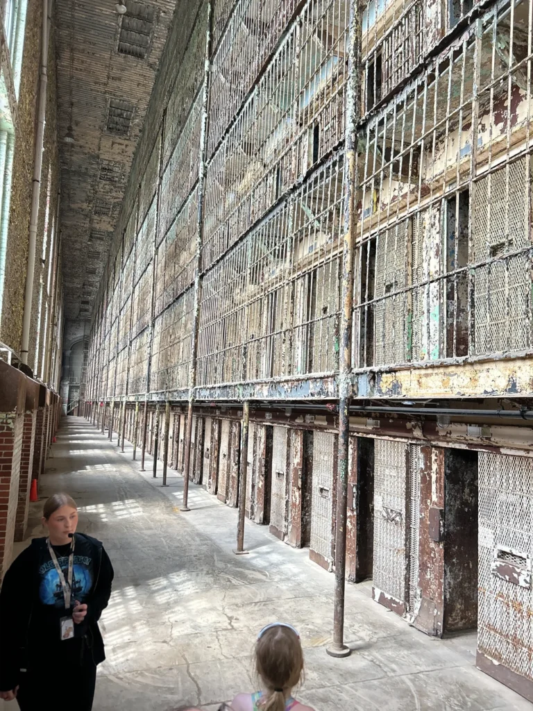 A towering, six-story steel structure filled with rows of small prison cells. The metal bars and walkways stretch upward toward a distant ceiling, creating a repetitive, industrial pattern of shadows and light. The towering six-story steel structure of the historic East Cell Block during a guided tour of the Ohio State Reformatory in Mansfield, Ohio.