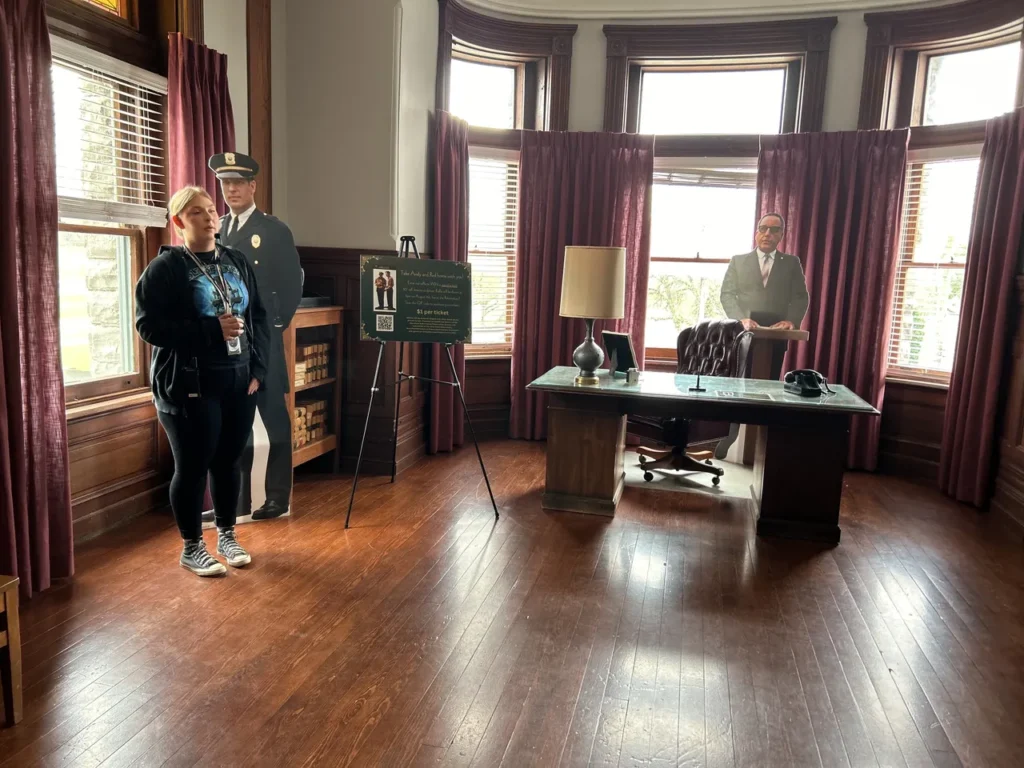 The Warden's Office at the Ohio State Reformatory featuring the dark wood desk, the 'His Judgment Cometh' embroidery, and the hidden wall safe used in The Shawshank Redemption. A group of three visitors posing on the grounds outside the historic Gothic stone castle entrance of the Ohio State Reformatory on a Shawshank Redemption prison tour.
