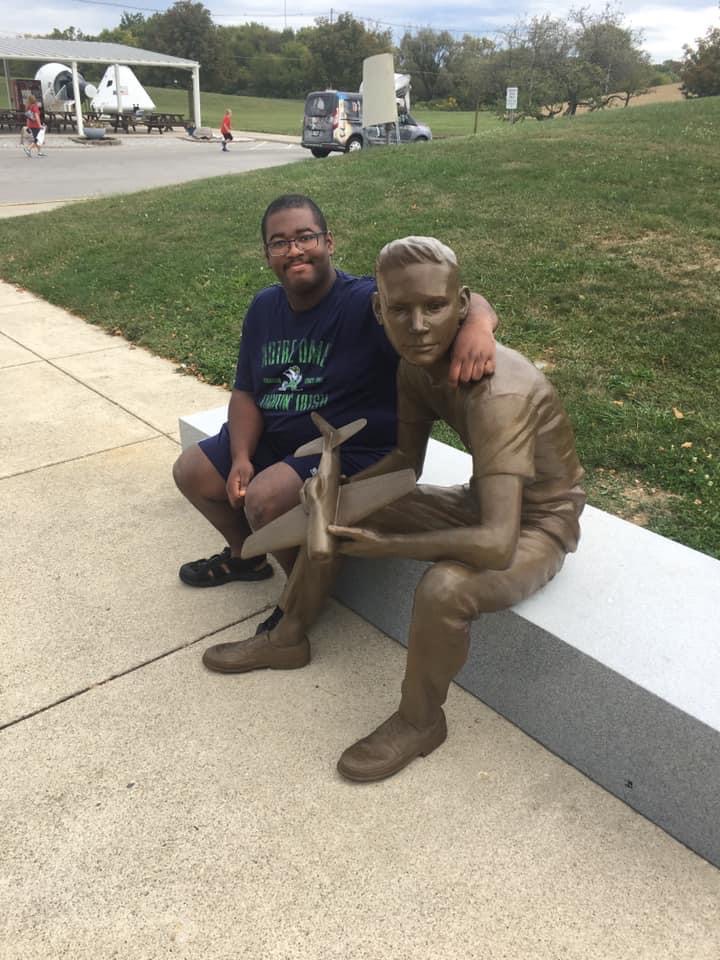A visitor sitting on a bench next to the bronze statue of a young Neil Armstrong holding a model airplane outside the Neil Armstrong Air and Space Museum in Wapakoneta, Ohio.